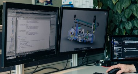 Zwei Monitore an einem Arbeitsplatz zeigen Software-Code und ein 3D-CAD-Modell einer Maschine.