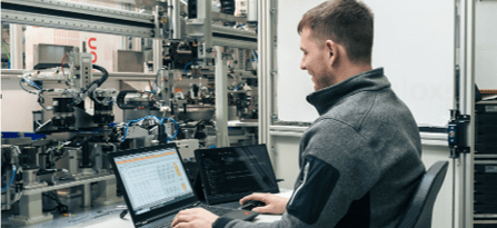 Techniker arbeitet an einem Laptop vor einer automatisierten Industrieanlage.